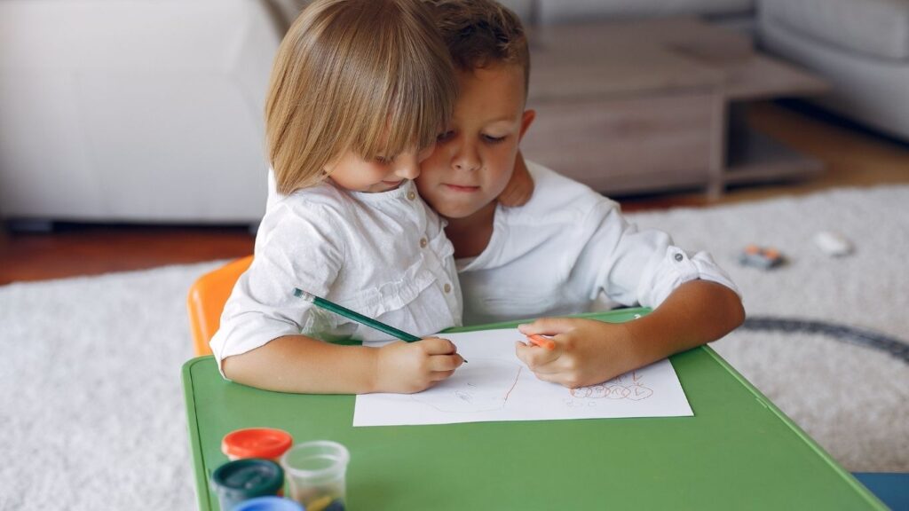 Two children drawing together with crayons at a green table in a bright living room